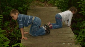 M and K on their knees looking over the edges of a boardwalk in the swamp